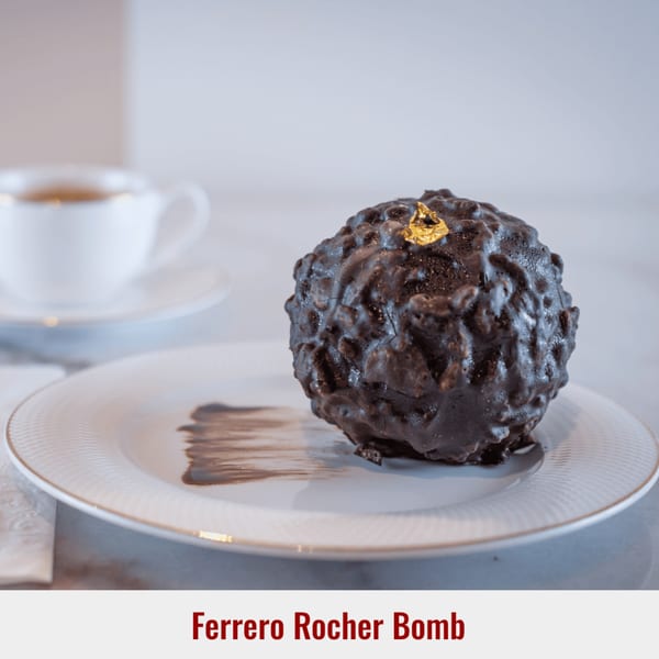 A Ferrero Rocher Bomb dessert, a chocolate sphere with gold leaf, on a white plate with a coffee cup.