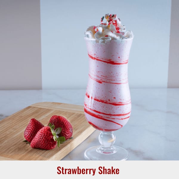 A strawberry shake with whipped cream and sprinkles, next to three fresh strawberries on a wooden board.