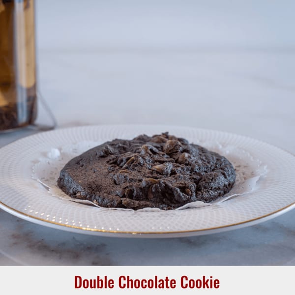A large, dark double chocolate cookie with chocolate chips sits on a white plate with a doily.