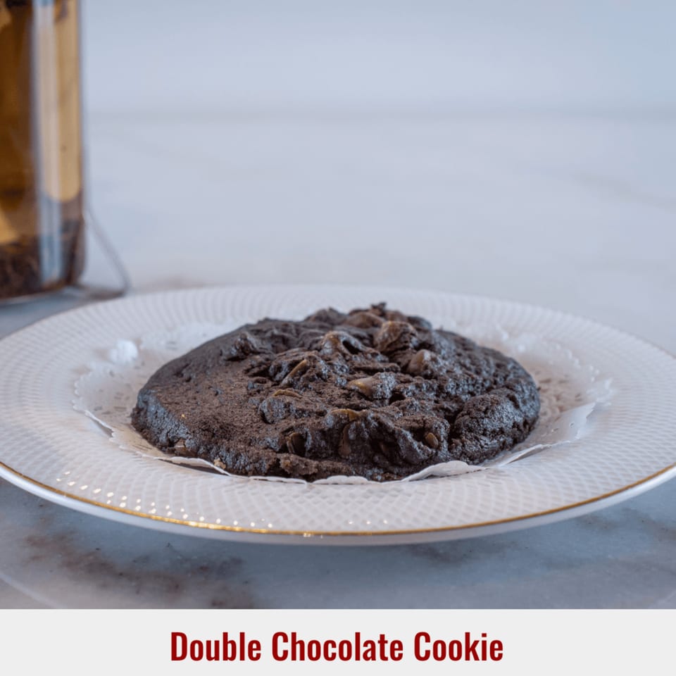 A large, dark double chocolate cookie with chocolate chips sits on a white plate with a doily.