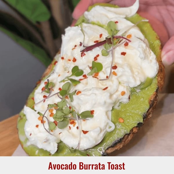Avocado Burrata Toast from Dessert Avenue, topped with microgreens and chili flakes, held by a hand.