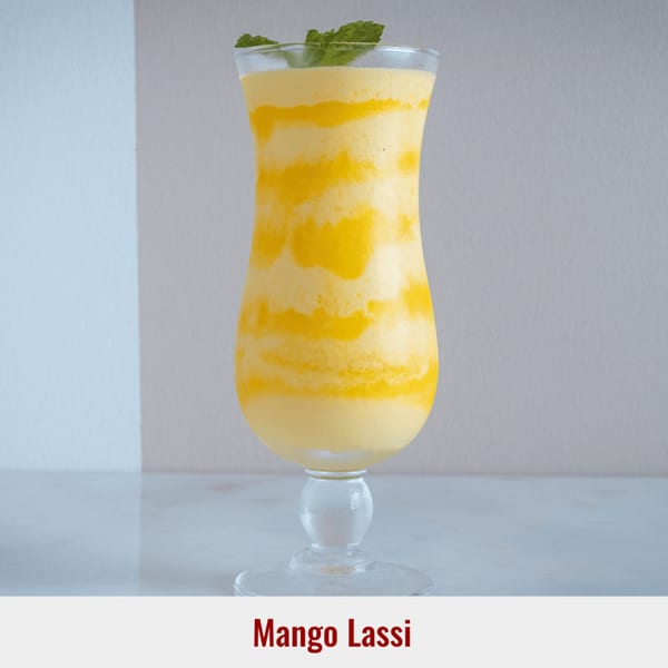A tall glass of Mango Lassi, a yellow and white swirled drink, garnished with mint leaves.