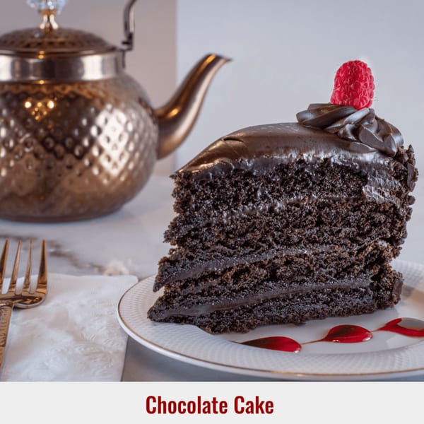 A slice of rich chocolate cake with frosting and a raspberry on top, next to a golden teapot.