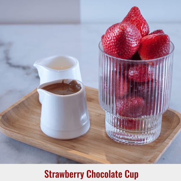Fresh strawberries in a glass cup with two small pitchers of chocolate and white sauce on a wooden tray.
