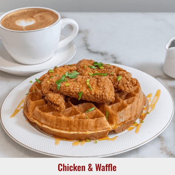 Golden-brown chicken and waffle dish with syrup drizzle and chopped chives, alongside a latte with heart art.