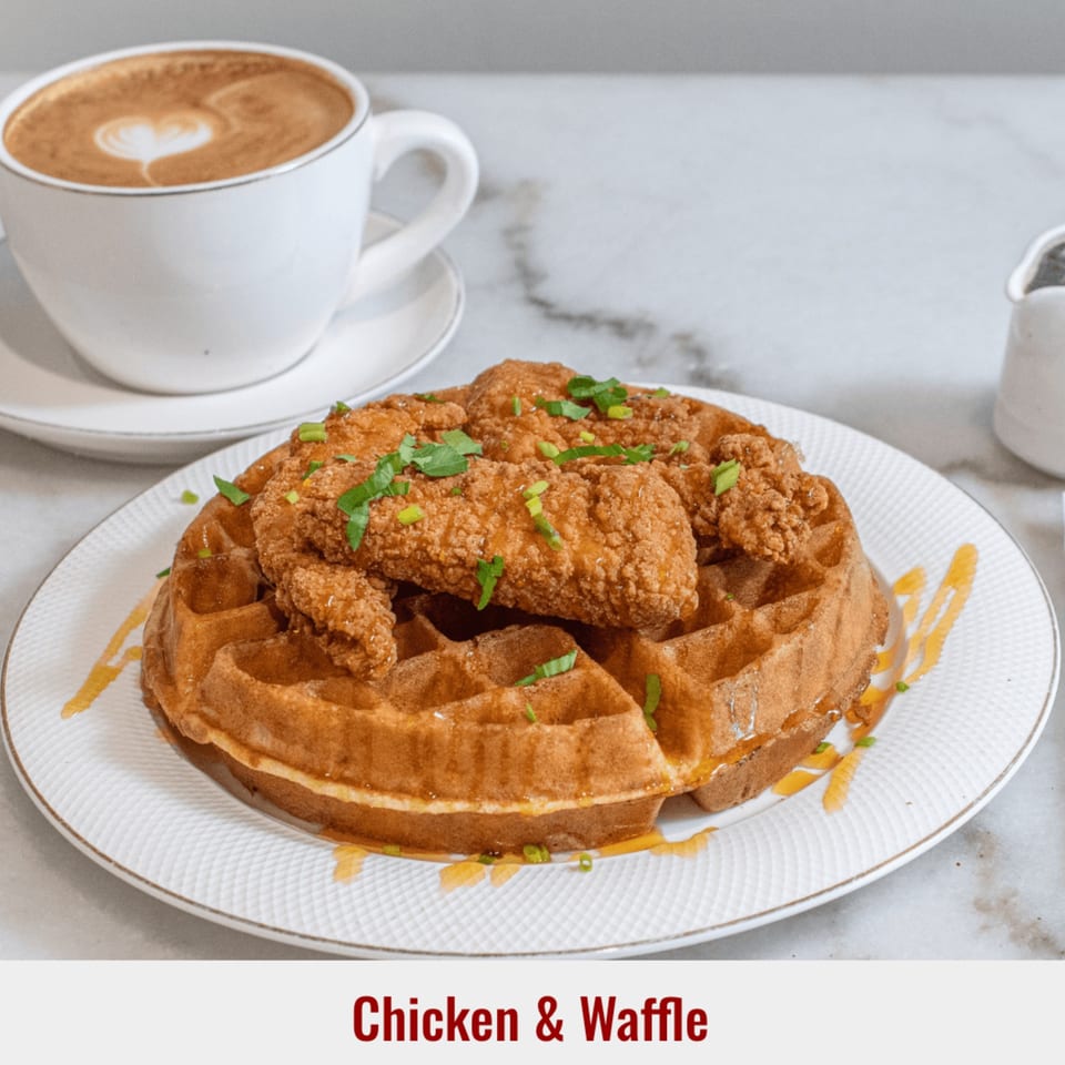 Golden-brown chicken and waffle dish with syrup drizzle and chopped chives, alongside a latte with heart art.