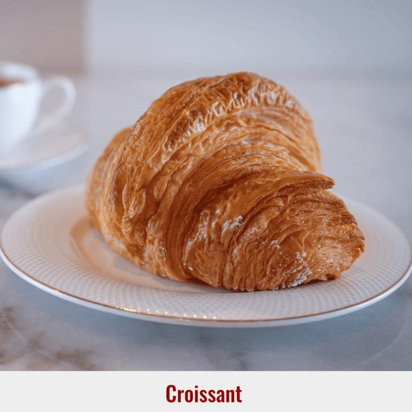 Golden-brown croissant on a white plate with a gold rim, with a coffee cup in the background.